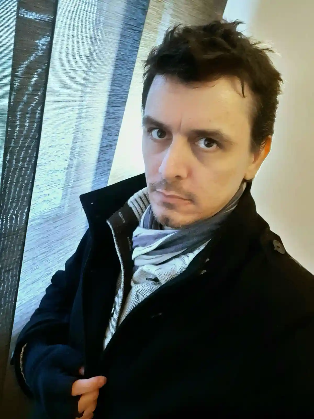 A man with dark, messy hair and a goatee stares intensely at the camera with wide eyes and a serious expression, wearing a…