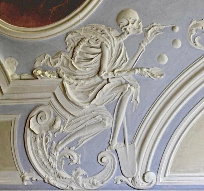 A white plaster relief sculpture of a skeleton dressed in draped robes, seated and playing billiards with three balls, set…