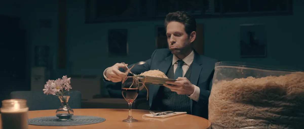 A man in a suit sits at a dimly lit dining table, looking disgusted as he tries to serve pasta from a plate into a wine…