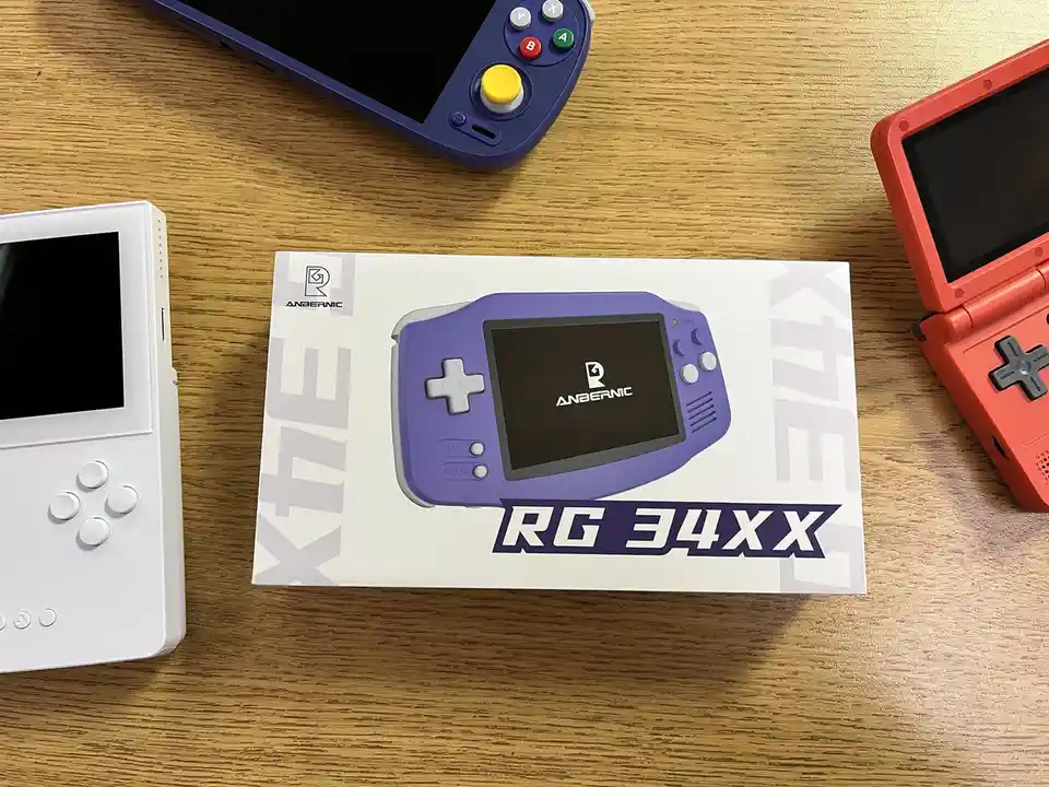 A white box for an Anbernic RG 34XX handheld gaming console sits on a wooden table, flanked by a white handheld, a blue…