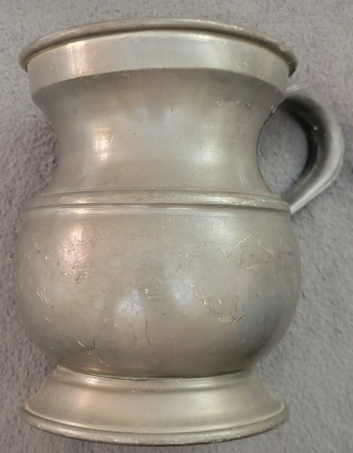 A close-up photo shows a silver-colored metal urinal with a handle on the right side, resting on a textured gray surface…