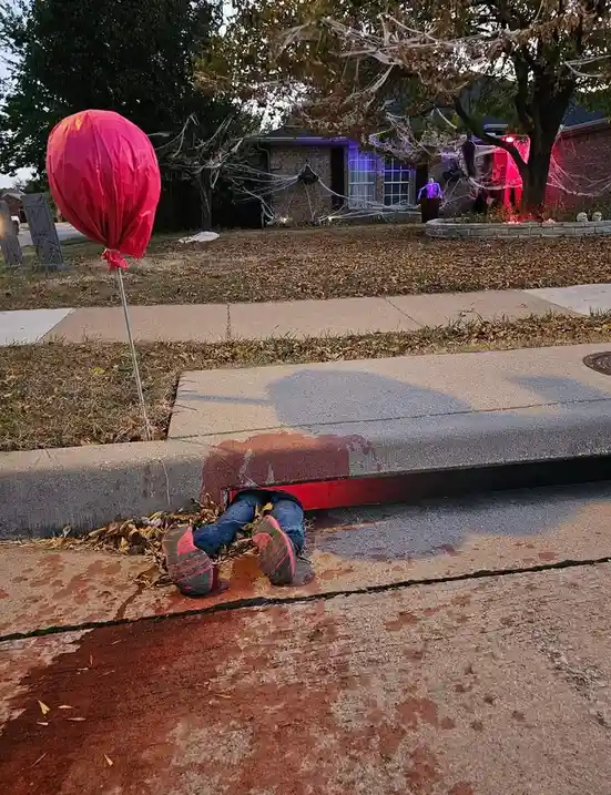 A person's legs and feet are visible sticking out from under a red Halloween decoration on a sidewalk curb, with fake blood…