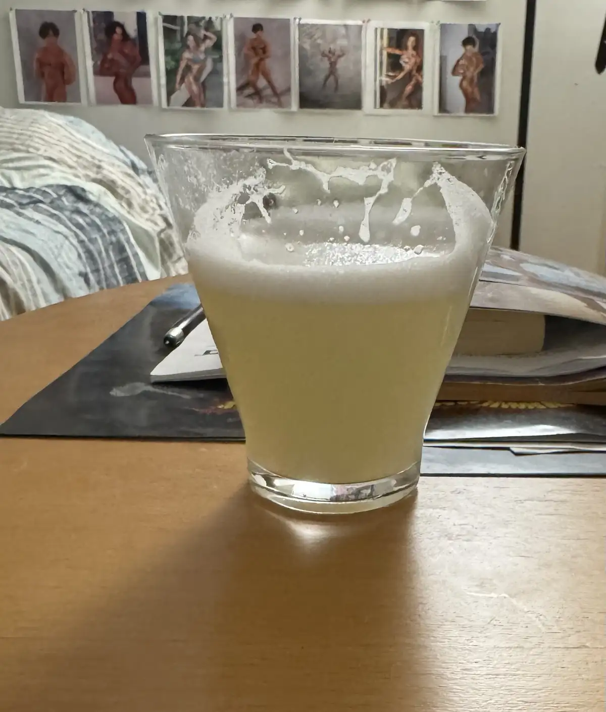 A glass of foamy, pale yellow beverage sits on a wooden table, with a row of framed nude art prints hanging on the wall…