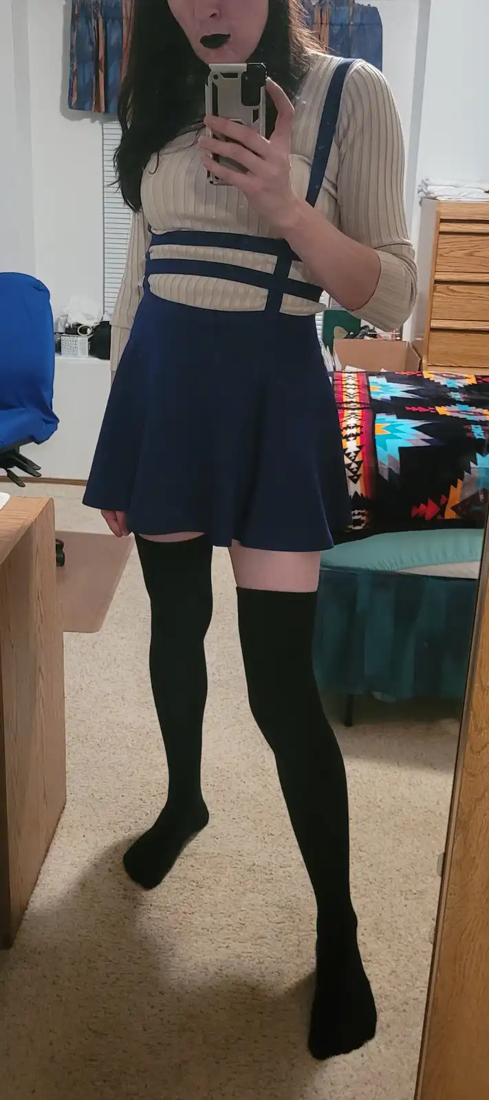 A person takes a mirror selfie in a bedroom wearing a beige ribbed top with navy blue suspenders and a matching navy blue…