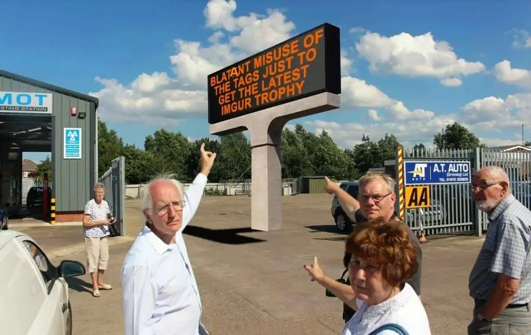 A group of older adults stand outside an MOT testing station, pointing at a large electronic sign that reads 'BLATANT MISUSE…