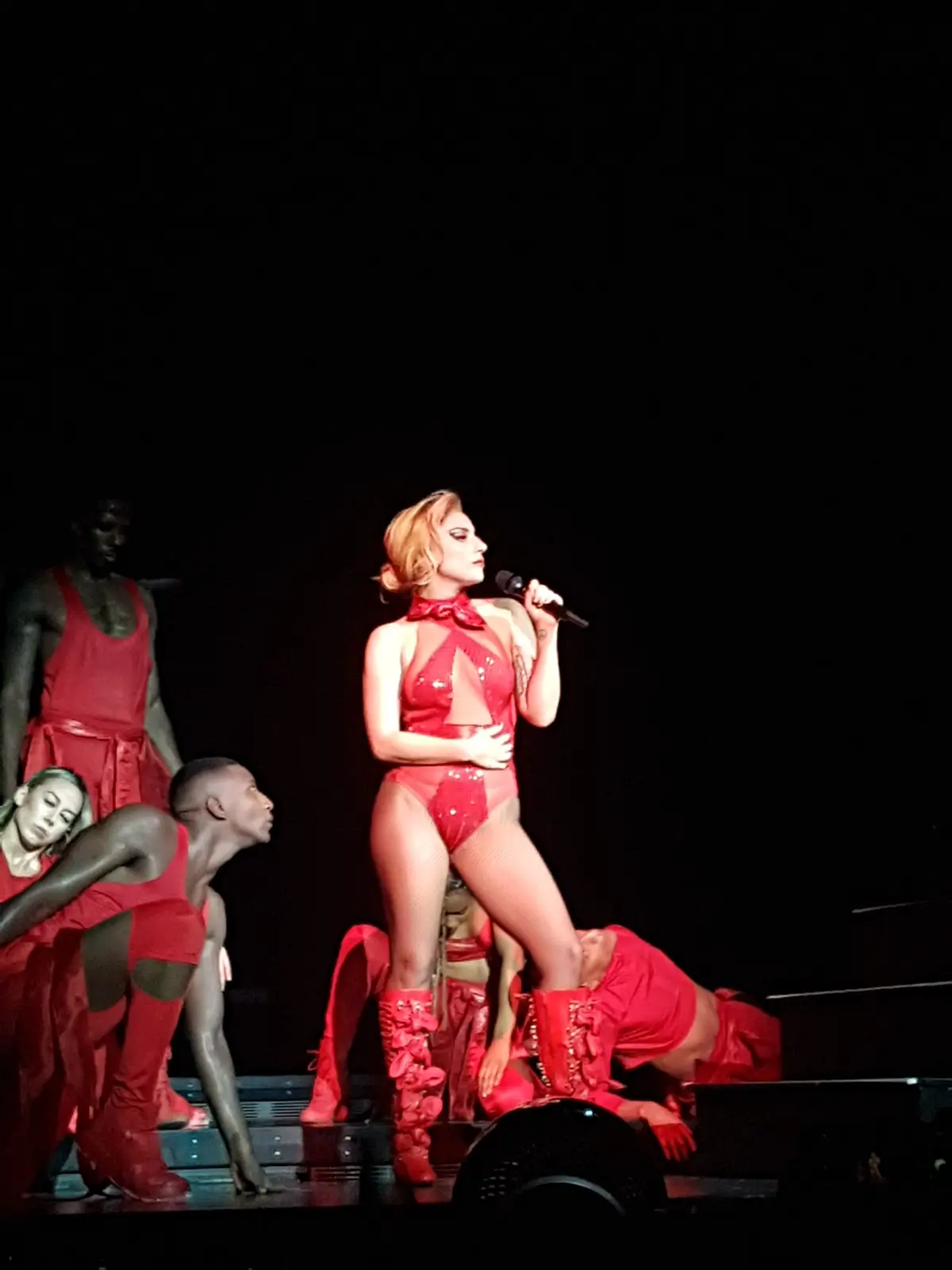 A woman in a red sequined outfit and tall red boots stands center stage holding a microphone, surrounded by dancers in red…