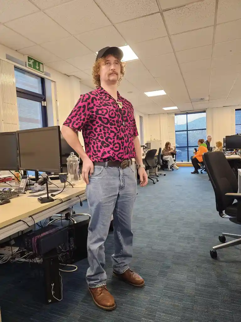 A man with curly hair and a mustache stands in an office wearing a bright pink leopard-print shirt, blue jeans, and a black…