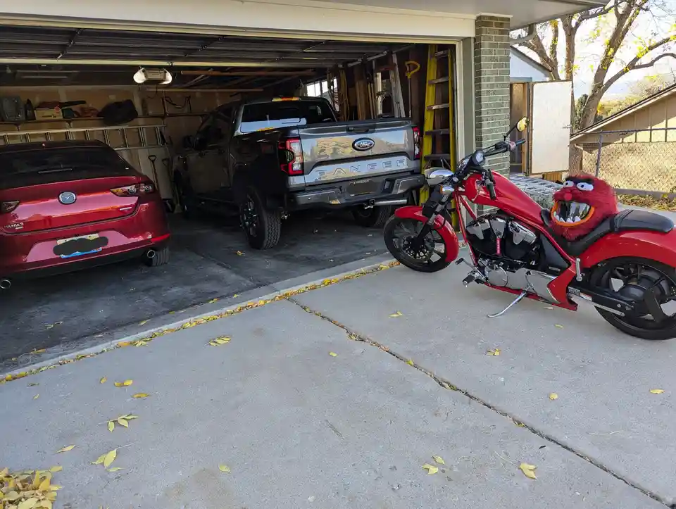 A red motorcycle with a red furry monster mask (resembling Animal from The Muppets) strapped to its seat sits parked outside…