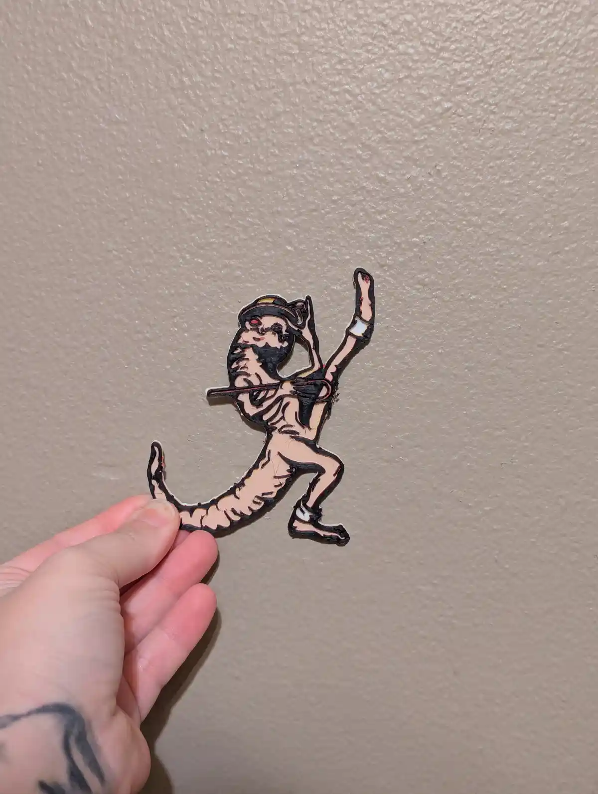 A hand holds up a cutout of a cartoonish, skeletal, tiger-like creature wearing a hat and holding a flute, against a beige…