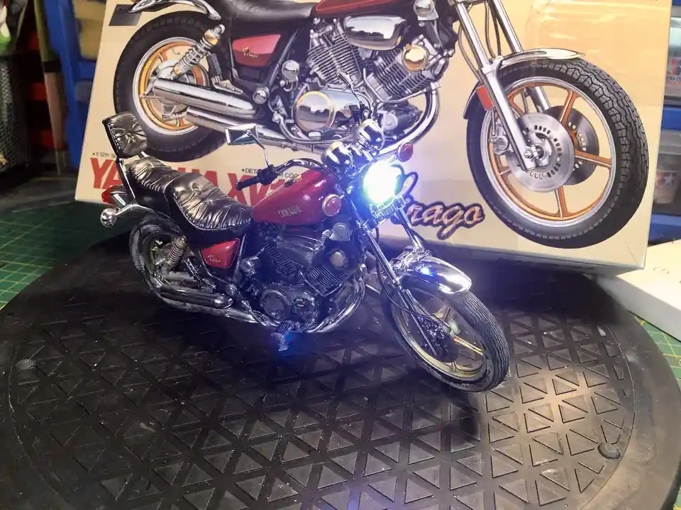 A highly detailed 1/12 scale model of a red and black Yamaha Virago motorcycle sits on a black textured display stand, its…