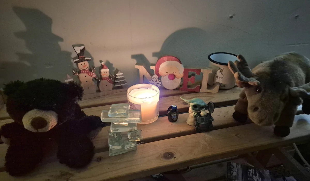 A dimly lit wooden shelf displays a collection of holiday decorations including snowmen, a Santa-themed 'NOEL' sign, a lit…