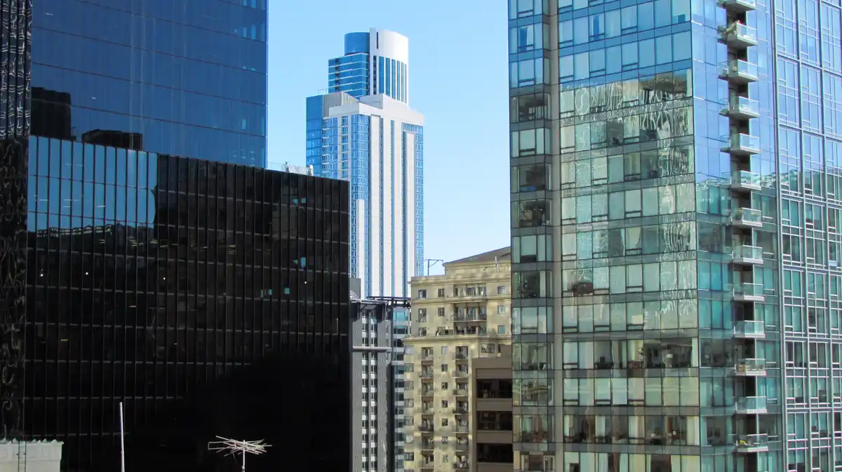 A cityscape photo shows several modern skyscrapers with reflective glass facades, including one with visible water streaks…