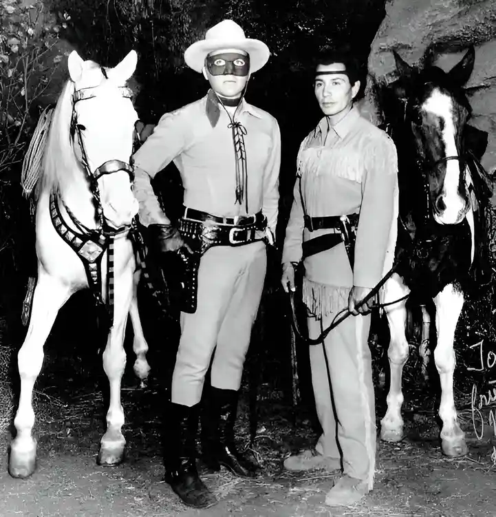 A black-and-white photo shows two men in Western attire standing beside horses. The man on the left wears a white cowboy hat…