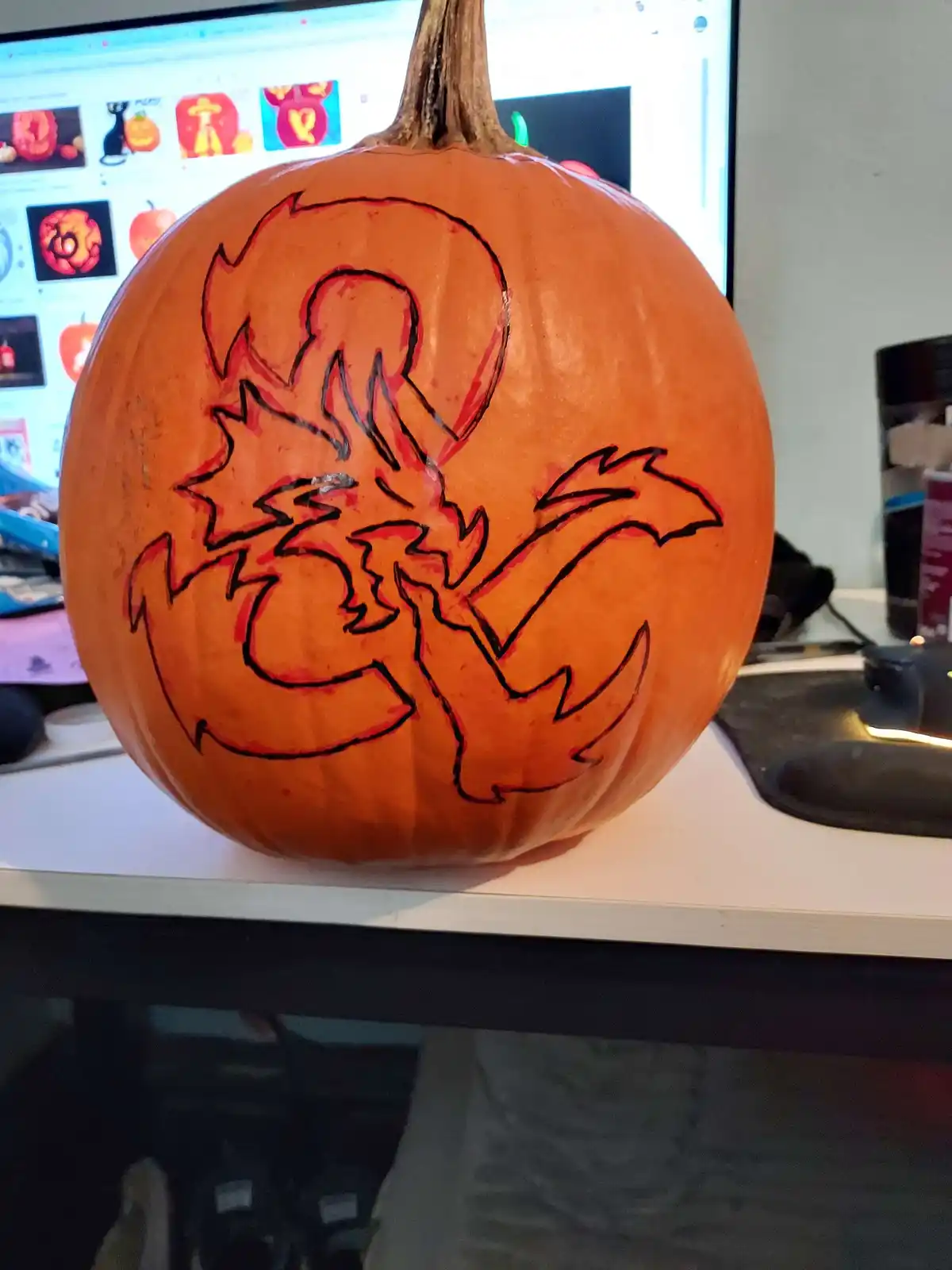 A close-up of an orange pumpkin sitting on a desk, with a crude black outline drawing of a dragon-like creature painted on…