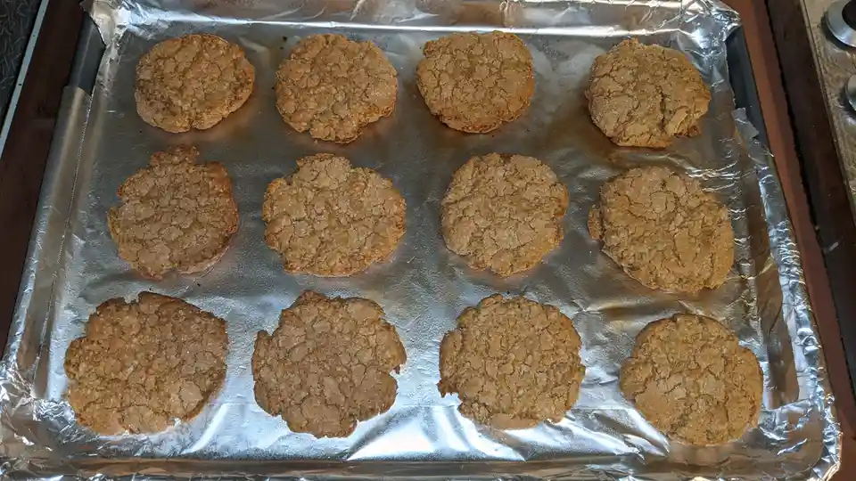 A baking sheet lined with aluminum foil holds twelve round, golden-brown cookies with a rough, cracked texture. The cookies…
