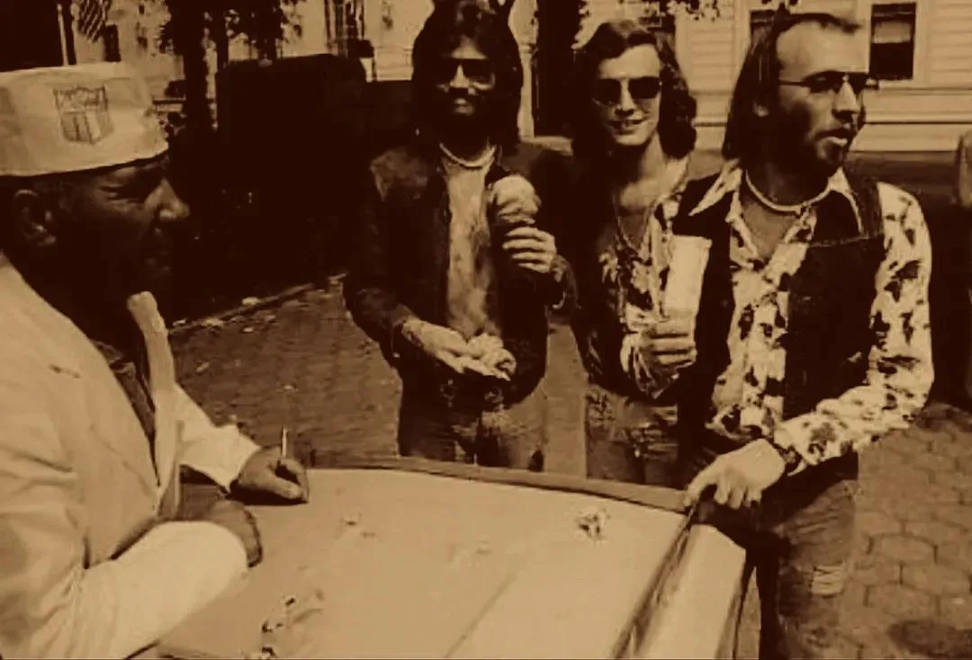 A sepia-toned photo shows a man in a white suit and cap signing a large, flat object while three men in 1970s-style clothing…