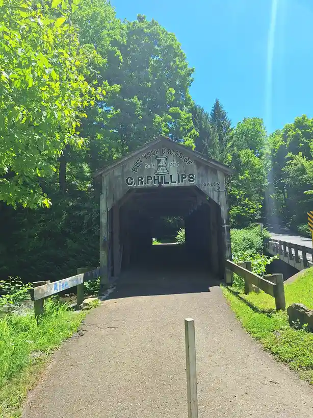 A wooden covered bridge with a faded sign reading 'BUY YOUR DRUGS OF C.R. PHILLIPS' and 'Built 1876' under a bright blue sky…