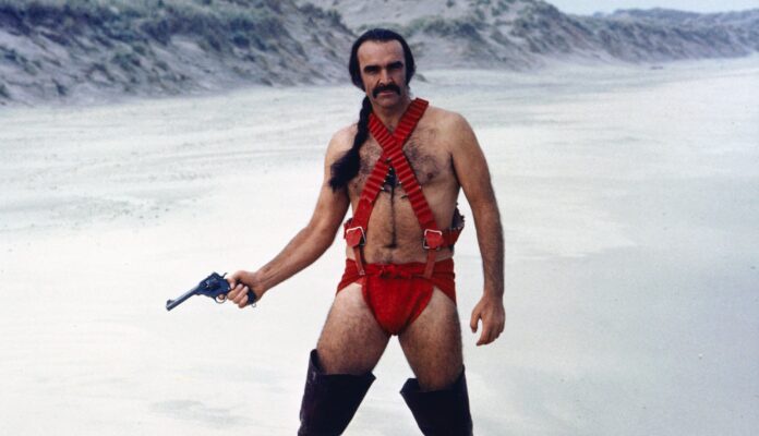 A man with a mustache and long dark hair in a ponytail stands in a desert-like landscape wearing red suspenders, red briefs…