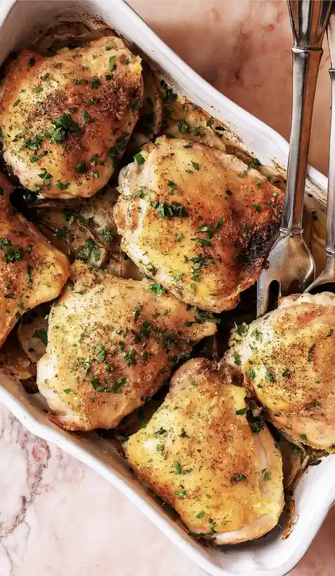 A close-up photo shows several golden-brown roasted chicken thighs nestled in a white ceramic baking dish, resting on sliced…