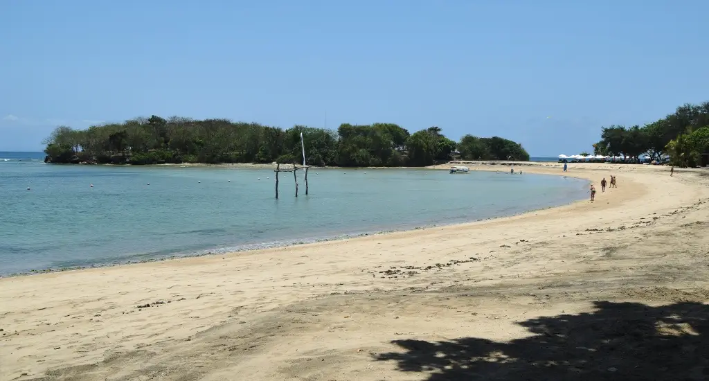 A wide-angle photo of a sunny beach with clear turquoise water, a sandy shore, and people walking in the distance. A wooden…