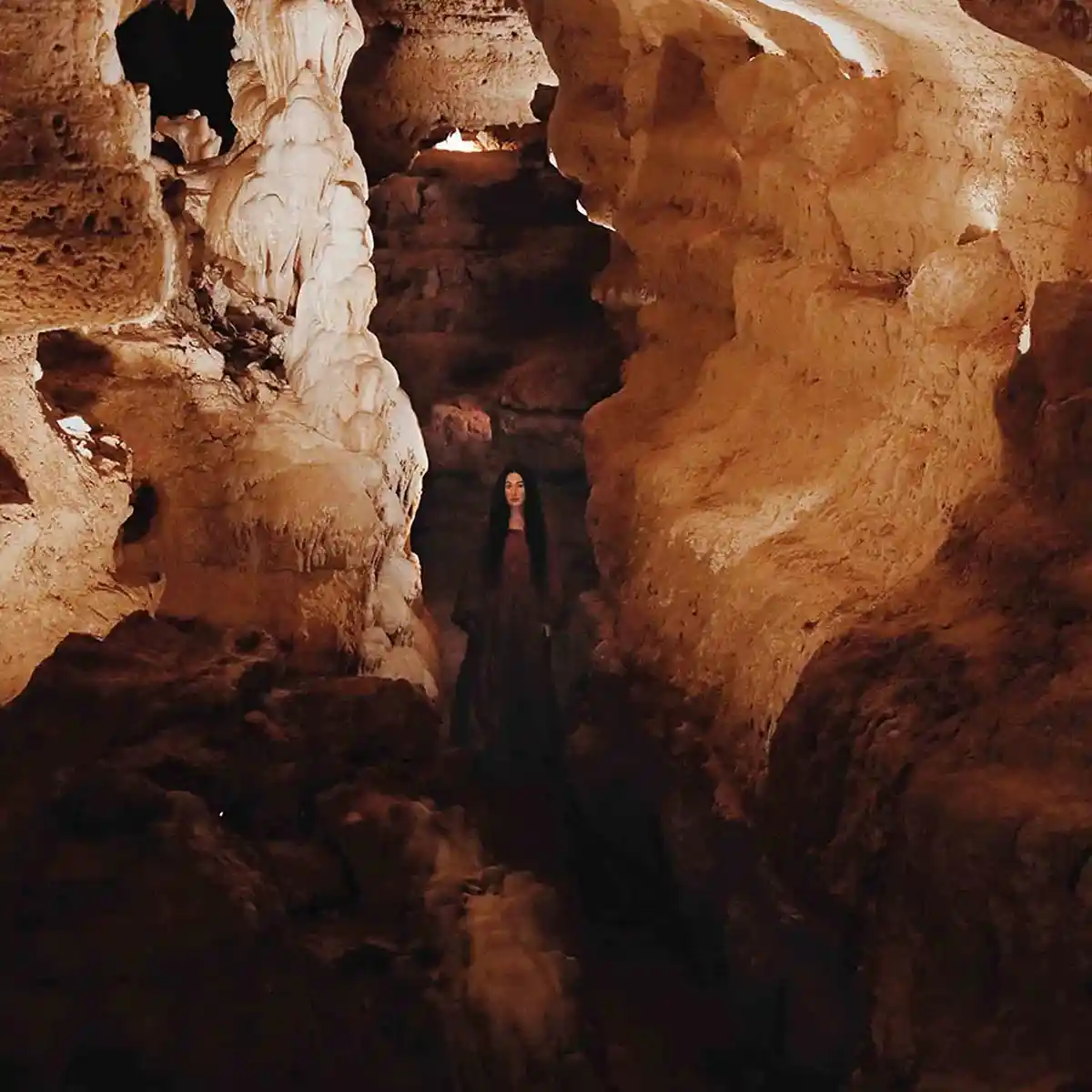 A woman with long dark hair wearing a dark robe stands in the center of a narrow, dimly lit cave with textured, orange-brown…