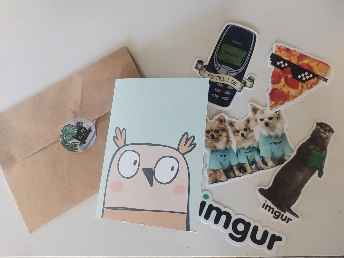 A flat lay of various Imgur-branded stickers on a white surface, including a cartoon owl on a card, a brown envelope with a…