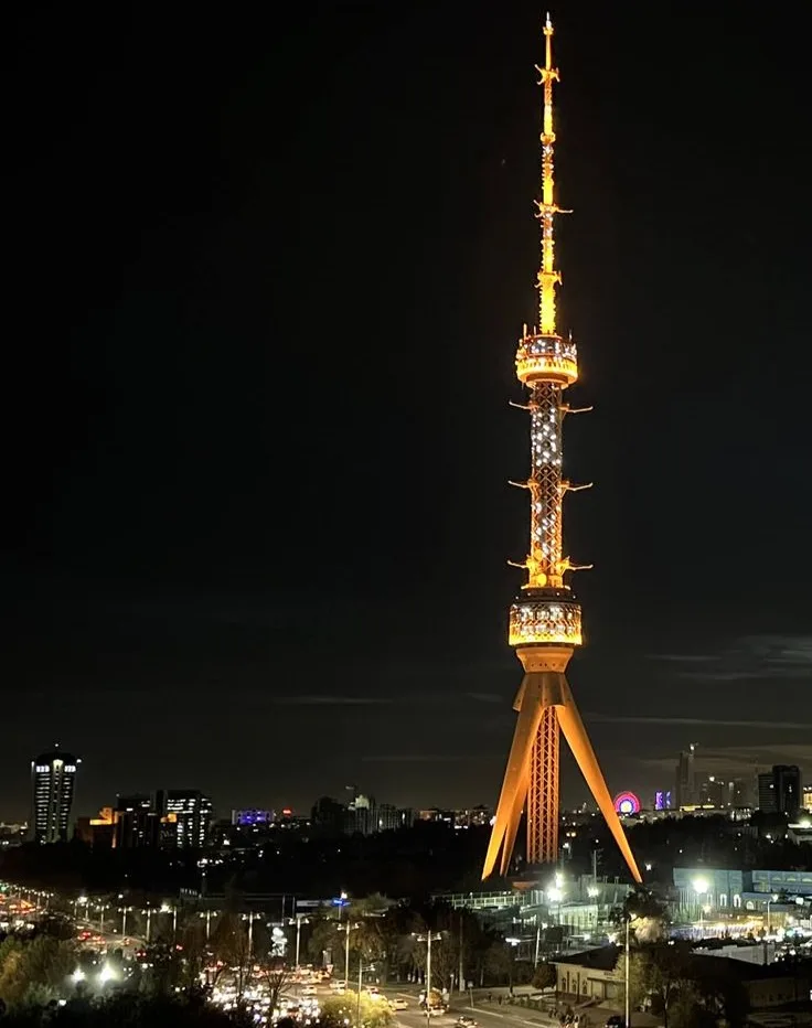 A tall, illuminated tower with a distinctive tripod base stands against a dark night sky, with city lights and buildings…