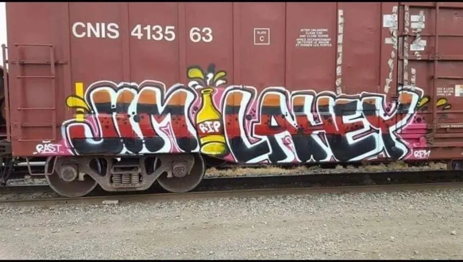 A red freight train car with graffiti reading 'RIP JIM LAHEY' in stylized letters, featuring a yellow bottle with 'RIP' on…