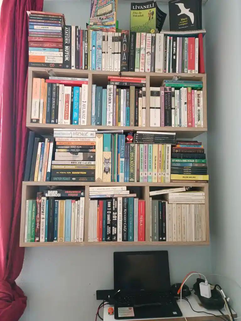 A wall-mounted wooden bookshelf is densely packed with books of various sizes and colors, mostly in Turkish, with titles…