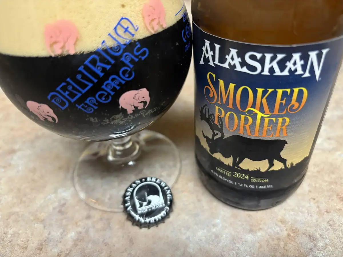 A close-up photo shows a bottle of Alaskan Smoked Porter next to a glass filled with the dark beer and topped with a thick…