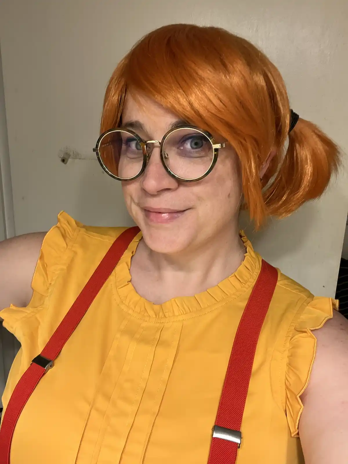 A person with orange pigtails, round gold-rimmed glasses, and a yellow ruffled top with red suspenders looks at the camera…