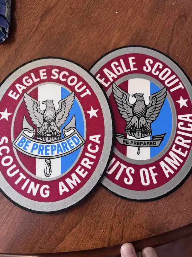 Two Eagle Scout patches from Scouting America lie on a wooden surface, one showing the standard 'BE PREPARED' banner and the…