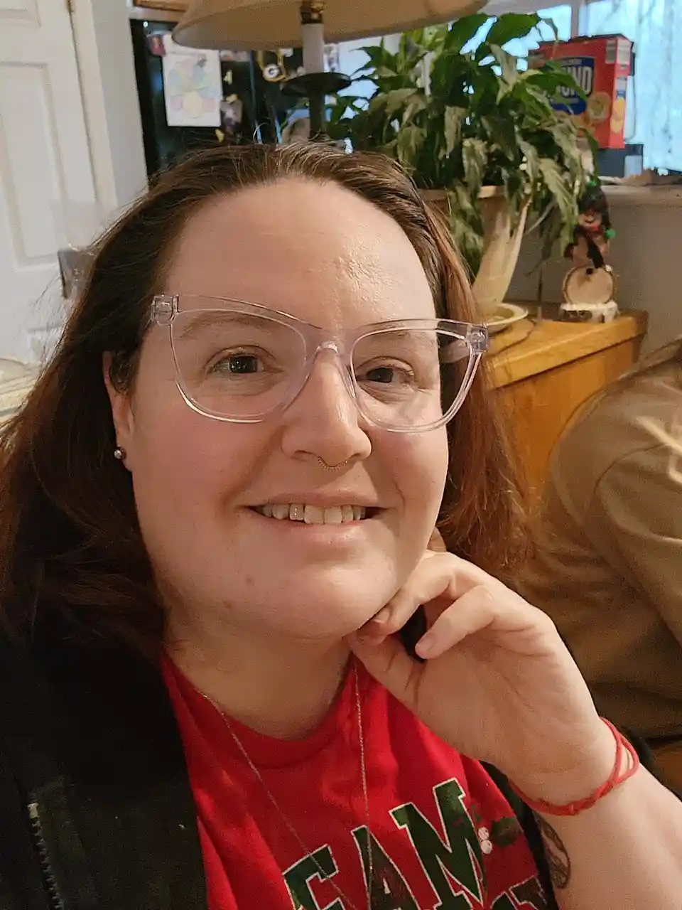 A woman with brown hair, clear-framed glasses, and a nose ring smiles while resting her chin on her hand. She wears a red…