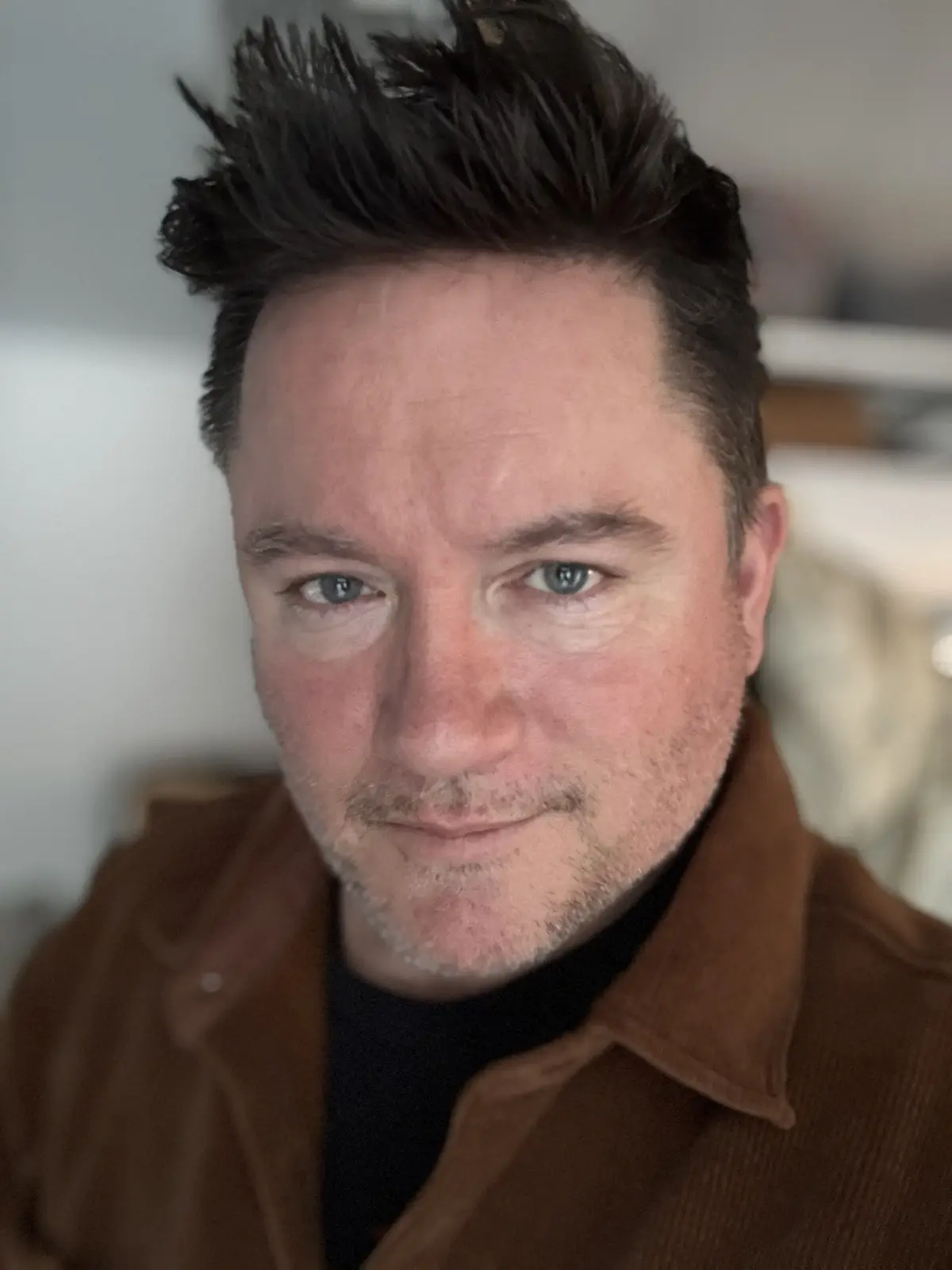 A close-up photo of a man with spiky, dark hair and light blue eyes, wearing a brown corduroy jacket over a black shirt…