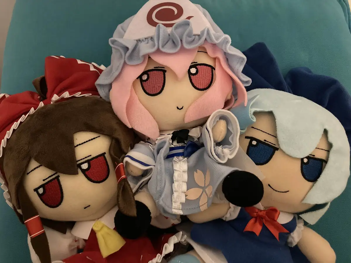 Three plush dolls of anime characters are huddled together on a teal couch. The central doll, with pink hair and a white…