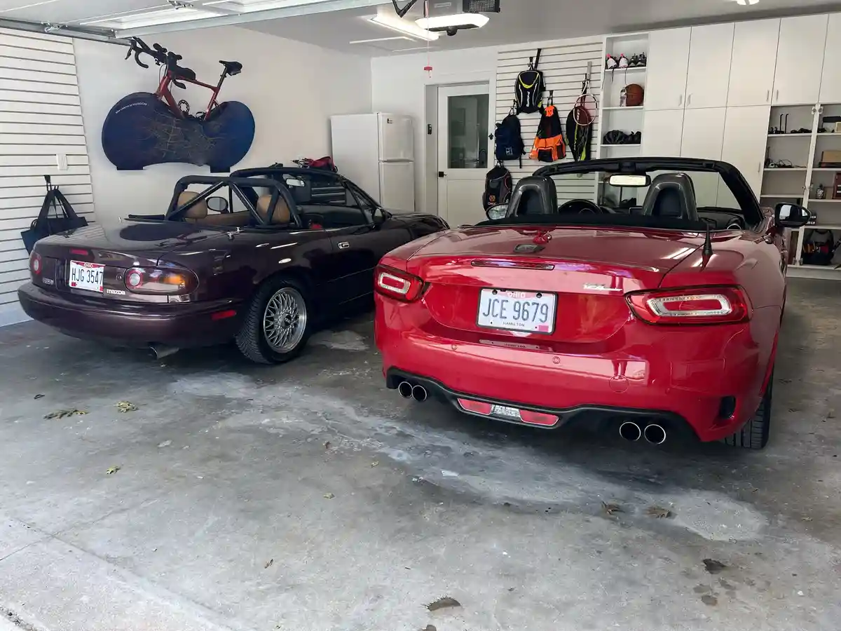 Two convertibles are parked side-by-side in a clean, organized garage. The red car on the right has a license plate reading…