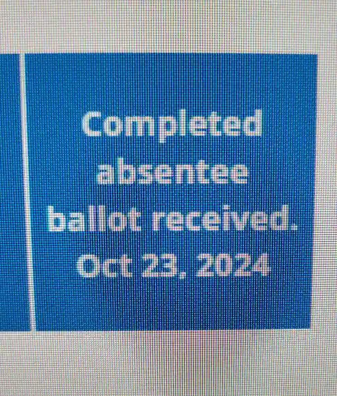 A close-up of a blue digital screen displaying white pixelated text that reads 'Completed absentee ballot received. Oct 23…