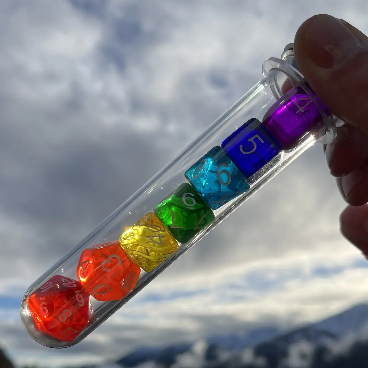 A hand holds a clear glass test tube against a cloudy sky with distant mountains, containing seven colorful, translucent…