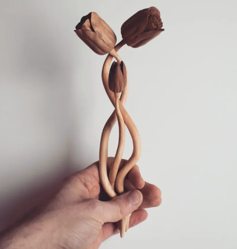 A hand holds a wooden sculpture of two intertwined flowers with three blooms total — two larger ones at the top resembling…