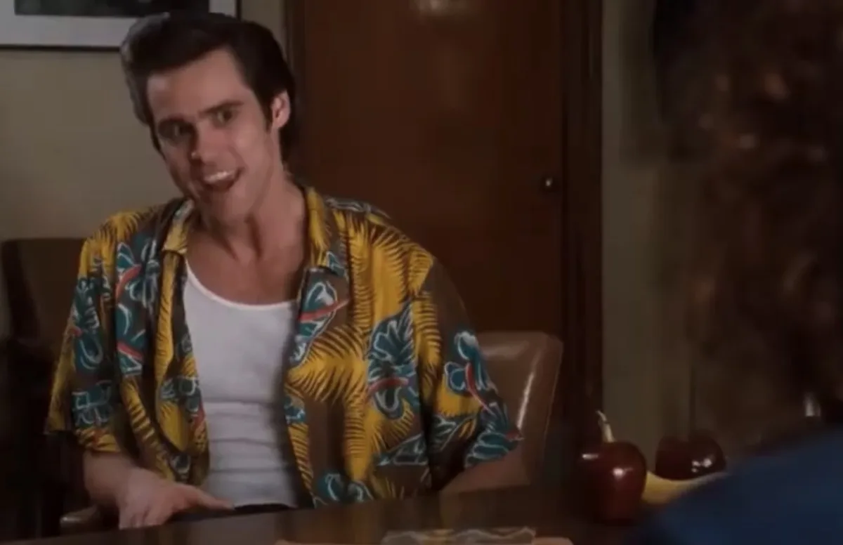 a man in a yellow and blue patterned shirt sits at a table, smiling broadly with wide eyes while looking at someone…