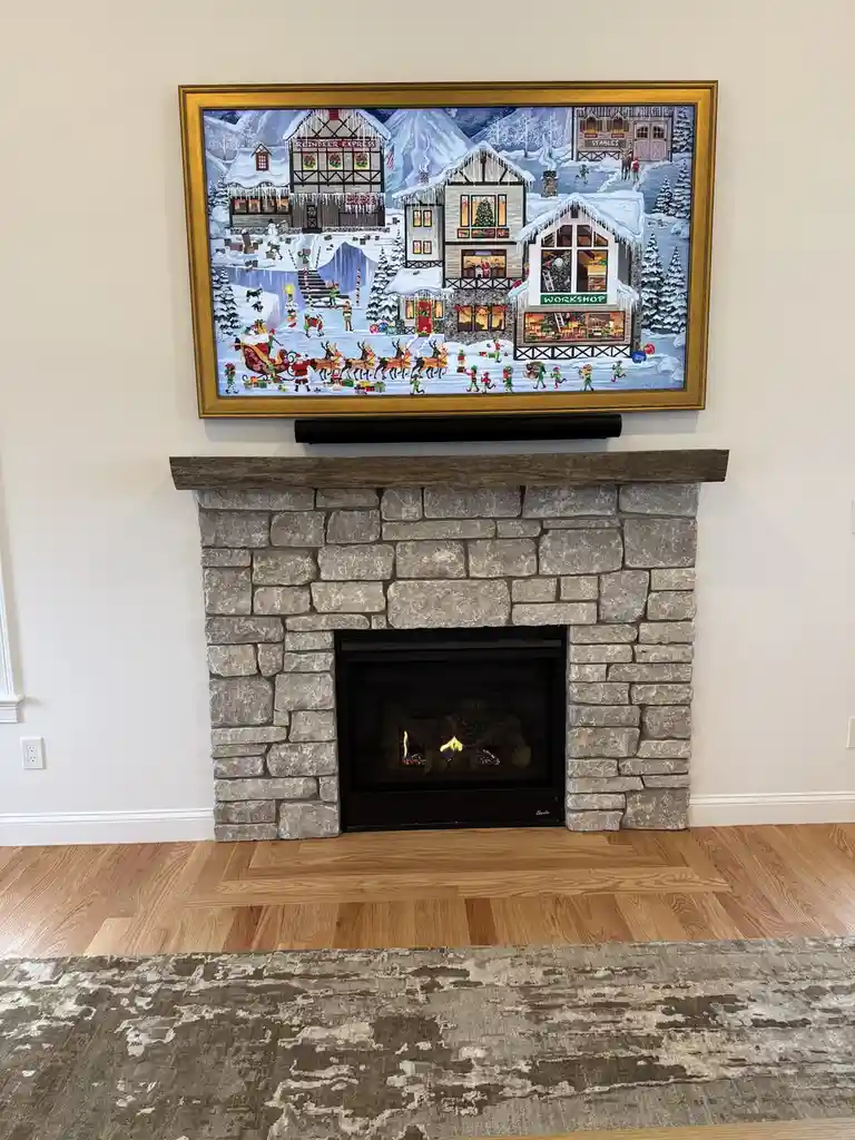 A stone fireplace with a lit fire is centered in a room with hardwood floors and a rug. Above the fireplace, a large framed…
