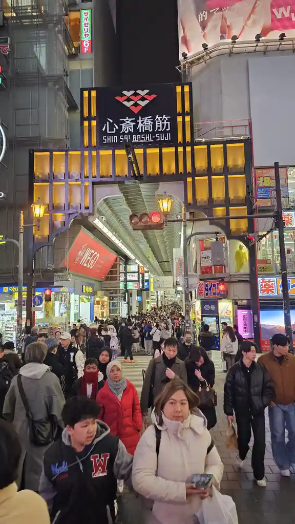 A crowded, brightly lit pedestrian street at night in Shin Saibashi-Suji, Osaka, Japan, filled with people walking under a…