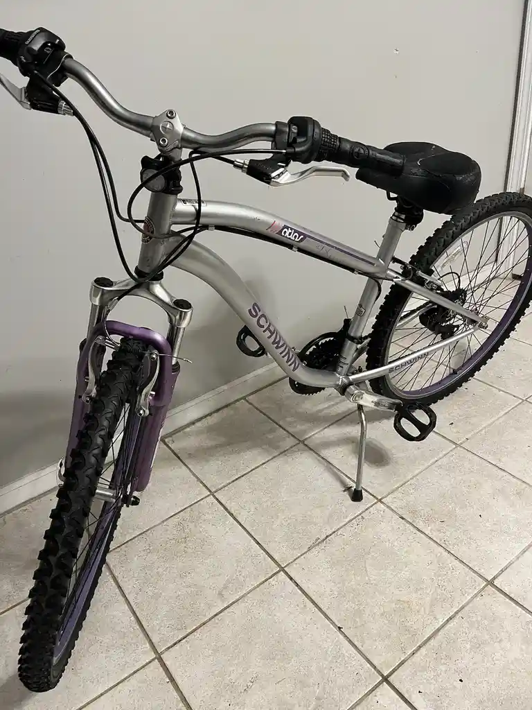 A silver Schwinn Atlas bicycle with purple accents is parked indoors on a tiled floor, its kickstand down and handlebars…