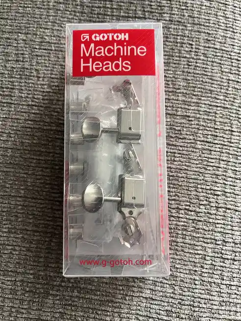 A clear plastic package of GOTOH brand machine heads for guitars, featuring shiny silver tuning pegs, sits on a gray…