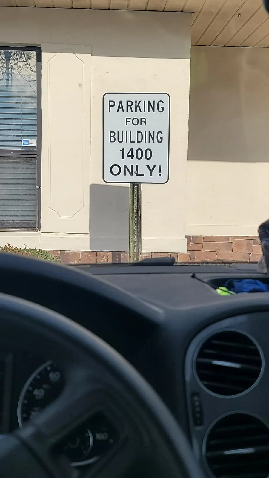 A photo taken from inside a car shows a white rectangular parking sign mounted on a metal pole, stating 'PARKING FOR…