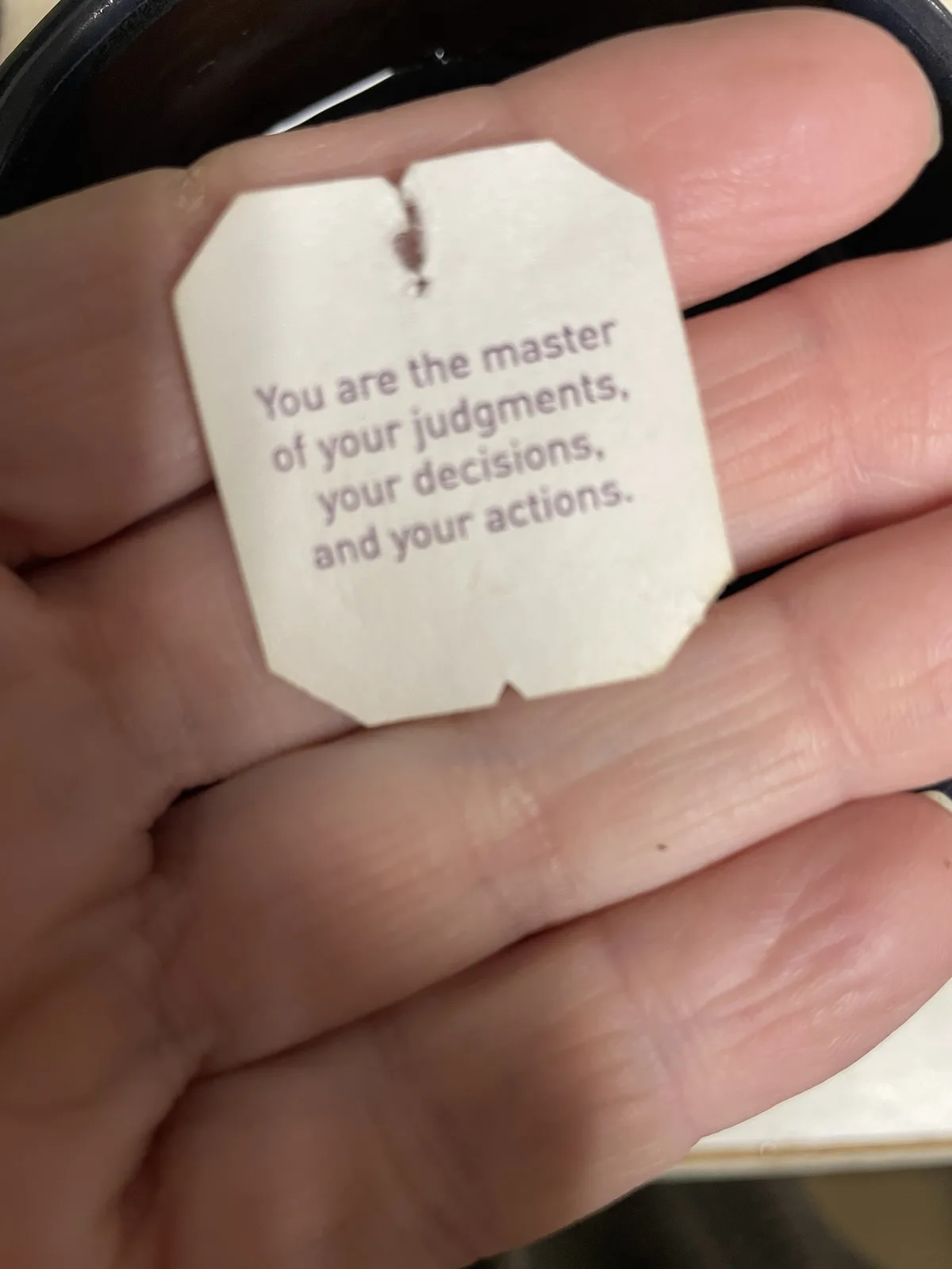 A close-up photo shows a person's hand holding a white, octagonal tea bag tag. The tag has purple text printed on it that…