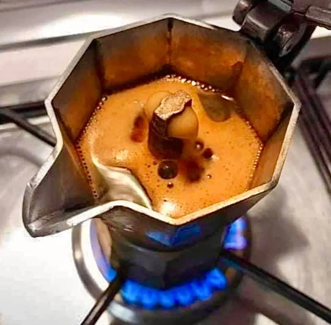 A close-up photo shows a metal moka pot on a gas stove with a blue flame underneath. Inside the pot, dark coffee is brewing…
