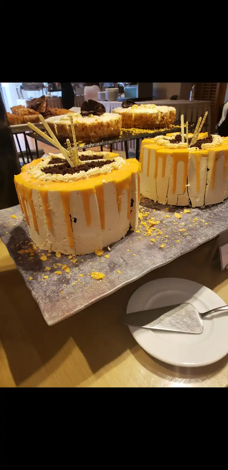 A photo of several large, sliced cakes with yellow sauce drizzled down the sides, displayed on a buffet table. A cake server…