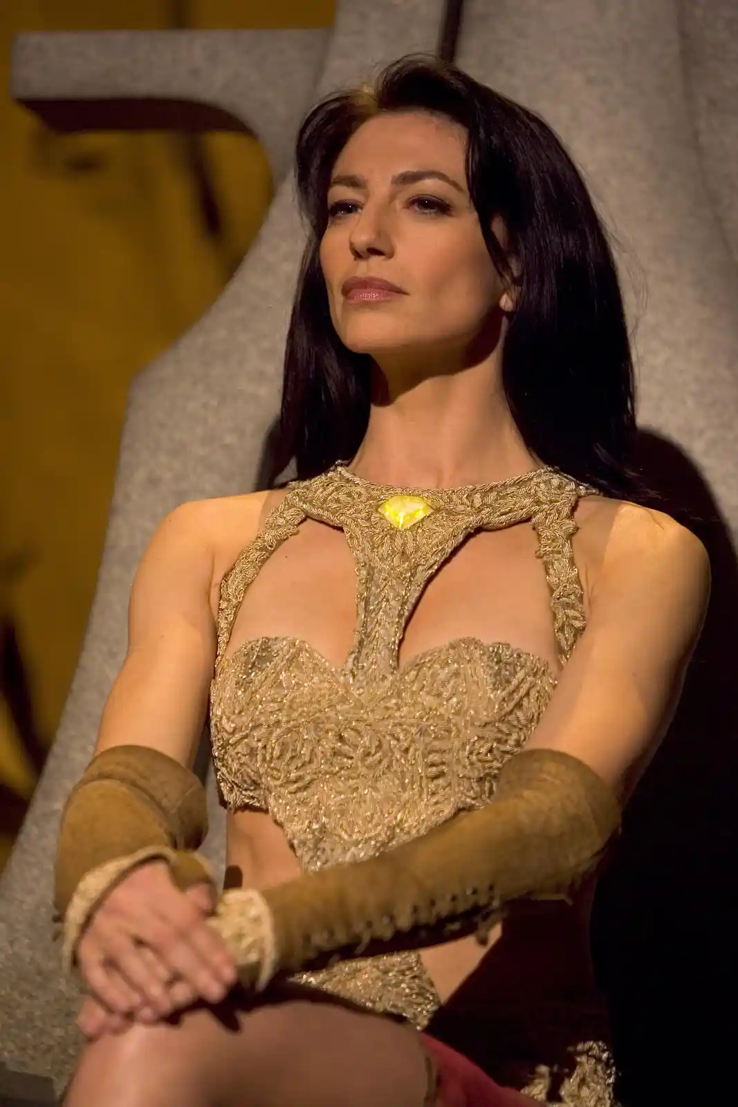 A woman with long dark hair sits in a grey chair wearing an ornate gold top with a central yellow gem and brown arm…