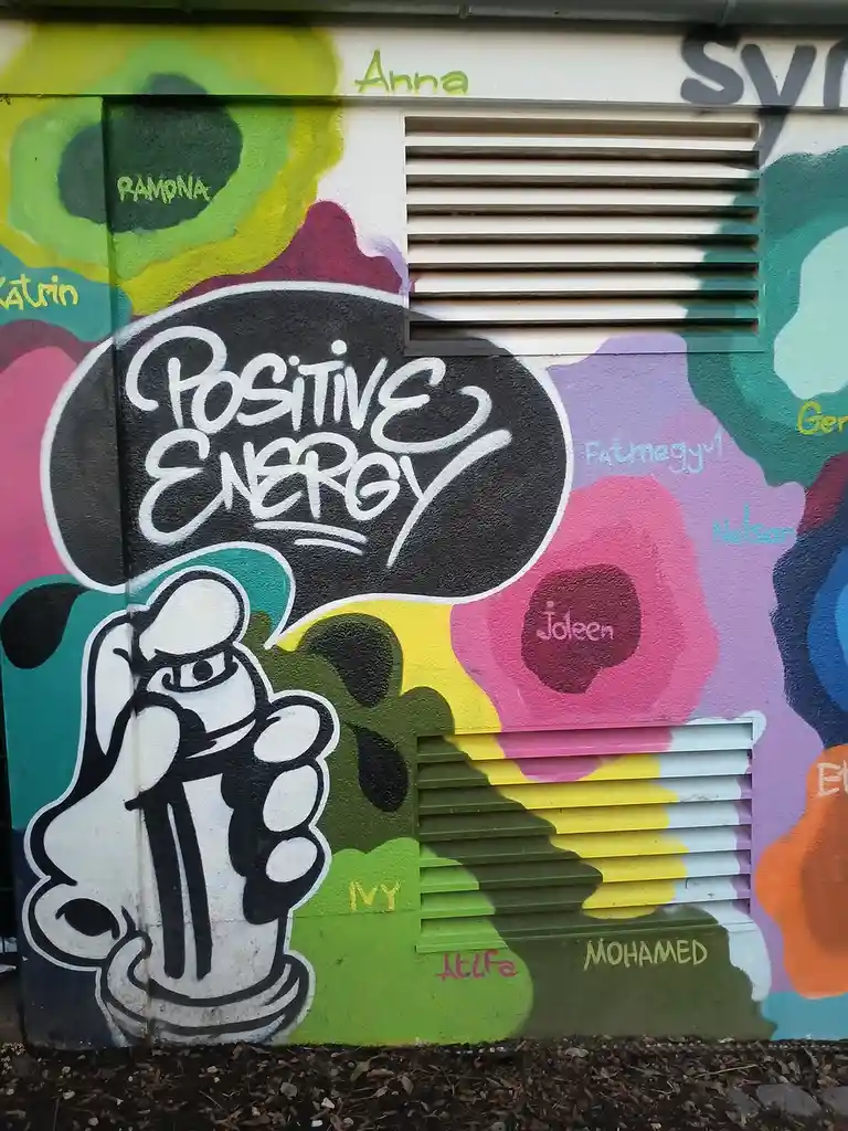 A colorful graffiti mural on a wall with two vents, featuring a cartoon fist holding a spray can and a large black speech…
